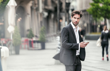 Attractive young businessman standing on the street and using his phone, windy weather