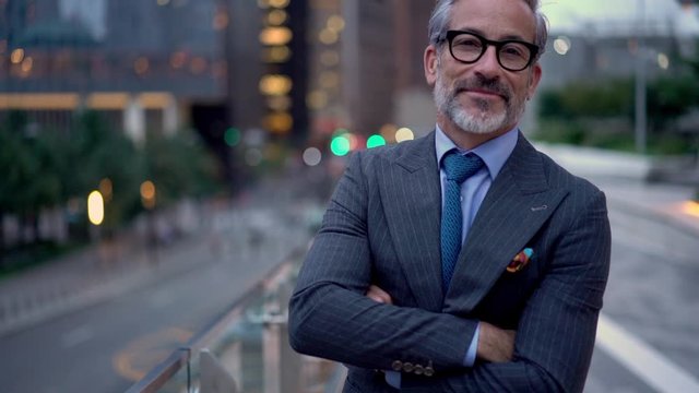 Slow motion of successful mature entrepreneur in expensive clothing posing in financial district with crossed hands and sincere smile on face, happy middle aged banker standing at urban setting 
