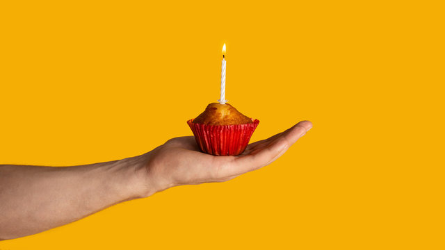 Festive occassion. Closeup of young guy holding tasty muffin with lit candle on orange background, panorama