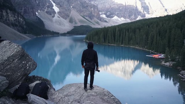 A Guy With Camera Standing On Top Of A Rock Looking At The Beautiful Moraine Lake In Banff National Park, Alberta, Canada - Descending Drone Shot