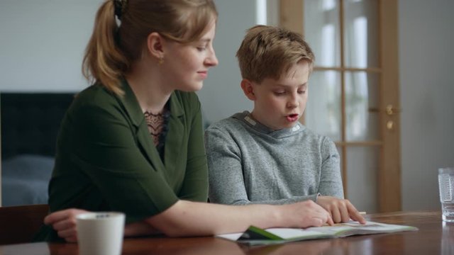 Big sister helps little brother with home work in home school. He points and writes in his book and they discuss the homework in a bright living room. He enjoys the home school lessons.