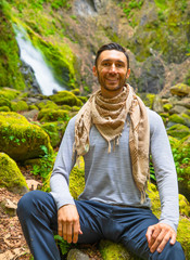 Man sitting in nature wearing scarf, long sleeve shirt and pants