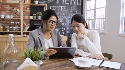 Young asian chinese businesswoman in suit explaining report to colleague at meeting in cafe store. two beautiful office lady coworkers sitting in coffee shop discussing project on digital tablet.