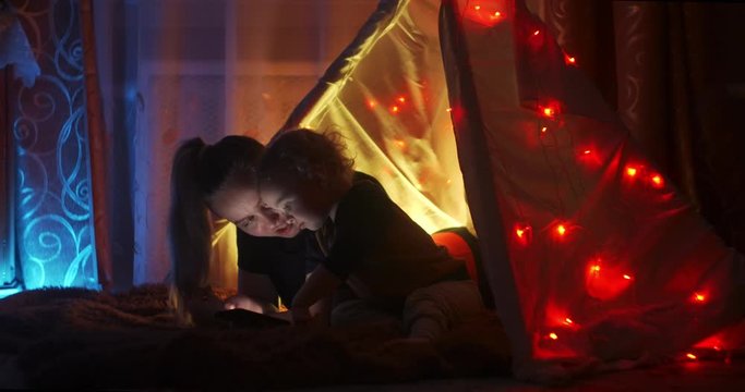 Bedtime story. Happy mother and little son read a book in a tent at home. Reading a book under a blanket with a flashlight in a dark room at night. Caucasian woman enjoying family time with her son.