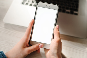 Woman's hands using empty white blank screen cell phone, working on laptop computer on table. Freelance. Work home.  Communication, technology concept. Girl checking social apps online. Top view.