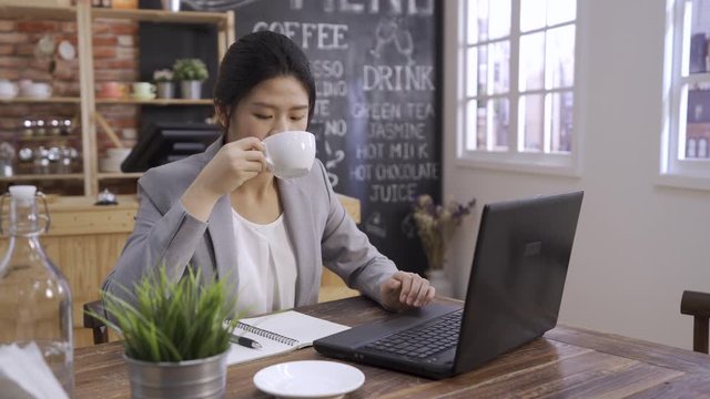 Young beautiful woman worker drinking coffee at cafe bar. elegant businesswoman in smart casual suit sitting at wooden desk enjoy hot drink in white cup while relaxing from working on laptop computer