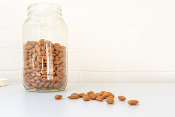 Close up of large glass jar of raw almonds on white table with handful scattered in foreground...