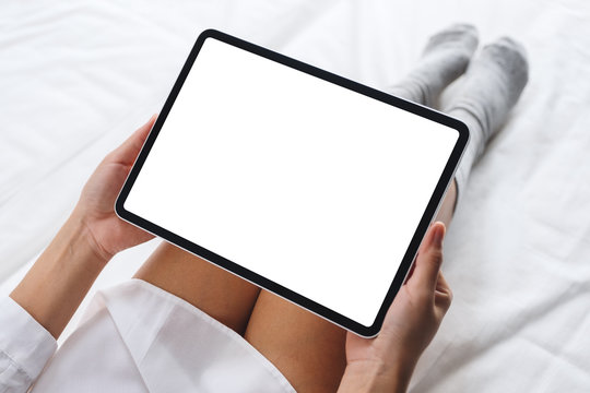 Top view mockup image of a woman holding black tablet pc with blank desktop white screen while sitting on a cozy white bed at home