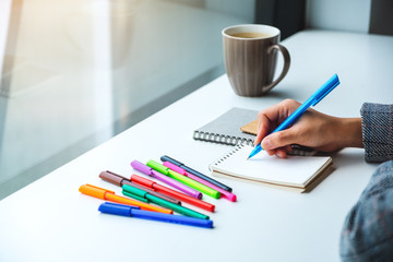 Closeup image of a woman writing on a blank notebook with colored pens and coffee cup on the table