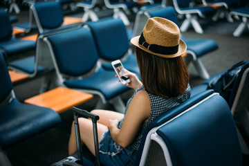 Young woman traveler sitting while using smartphone on chair of passenger in an airport lounge...