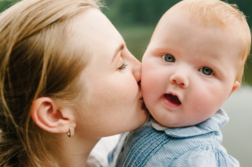Portrait mom kissing child together on nature. Mum, little daughter outdoors. Young mother with baby girl walk on beach near lake. Family holiday on pond. Happy Mothers Day. Close up.