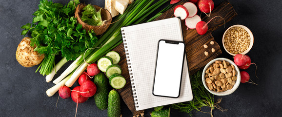 Order food online concept. Smartphone with blank screen and notepad with ingredients for cooking vegan food. Fresh vegetables, herbs, cereal and nuts on dark background top view