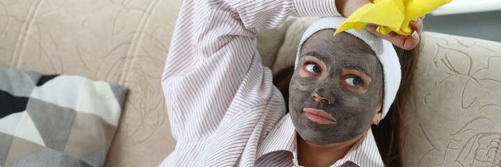 Portrait of tired young woman applying refreshing facial mask and sitting on couch. Pretty female holding yellow napkin to clean-up at home. Housewife and beauty concept