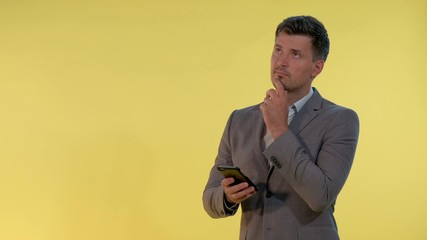 Young businessman in suit looking on smartphone and thinking about something on yellow background....