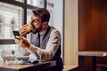 Hungry businessman sitting on lunch break in fast food restaurant, eating cheese burger and using...
