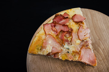 slice of tasty pizza on a wooden board on a black background. View from above. Pizza with ham and cheese. One piece. Copy space.