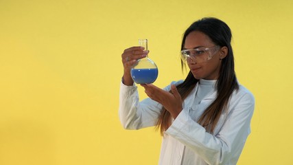 Pretty black woman in safety glasses and lab coat holding flask with liquid in her hands. There is yellow background.