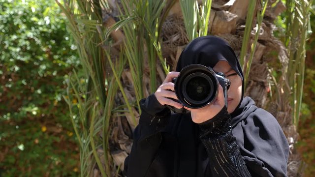 A beautiful Moroccan Arab Muslim woman with a DSLR camera filming photographing in a garden - turning the camera away from the viewer. Islamic emancipation. Real-time footage.