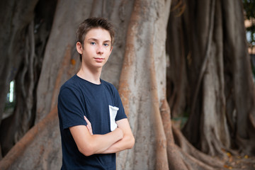 Young boy posing in summer park with ficus trees. Cute spectacled smiling happy teen boy 13 years old, looking at camera. Kid's outdoor portrait.