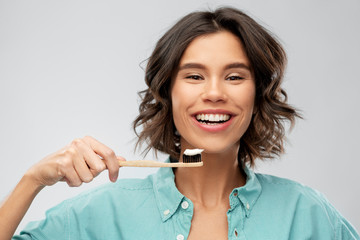 dental care, oral hygiene and people concept - portrait of happy smiling young woman in turquoise shirt with toothpaste on wooden toothbrush over gray background