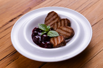 Grilled meat pieces with berry sauce served on a white plate over dark rustic wooden background. Italian cuisine.