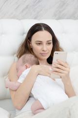 Mother and child on a white bed. Mom and baby boy playing in bedroom. Mother is using mobile phone on bed.