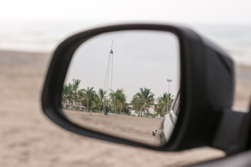 The side mirror of a car captures a serene view of palm trees and a distant beach, evoking a sense...