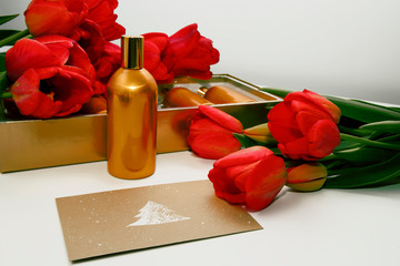 An envelope with a picture of spruce lies on the surface where there are a golden bottle and red tulips.