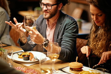 Hipster sitting with his friends at restaurant for diner, drinking wine and chatting.