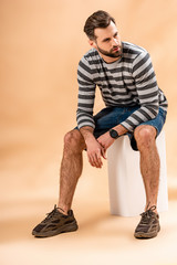 Naklejka premium bearded man in striped sweatshirt sitting on white cube on beige