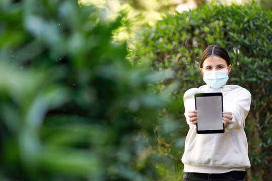 bambina con i capelli neri indossa una mascherina facciale per proteggersi e mostra un tablet al parco 