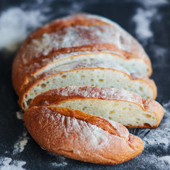 Delicious homemade wheat bread, cut into pieces, lies on a black tray strewn with flour. Home cooking. Bakery products.