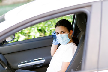 bambina con i capelli neri indossa una mascherina facciale e guanti in lattice per proteggersi e telefona dall'abitacolo di un automobile 