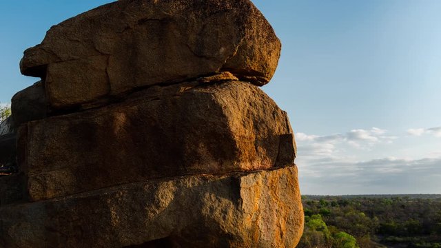Late afternoon linear timelapse of big granite rock boulders in golden sunlight showing texture, revealing African bush landscape in distance with green trees dramatic clouds moving, dip to black.