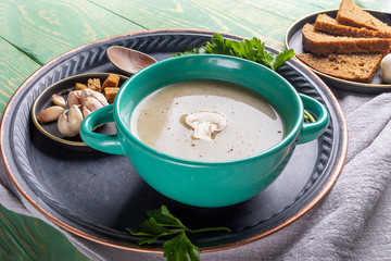 Close-up turquoise ceramic bowl with mushroom cream soup on round metal tray over green wooden background.