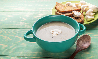 Turquoise ceramic bowl with mushroom cream soup, wooden spoon, garlic, bread, on green wooden background.