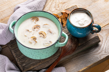 Turquoise ceramic pan with mushroom cream soup, cup with sour cream and crackers on old wooden board on wooden rustic background.