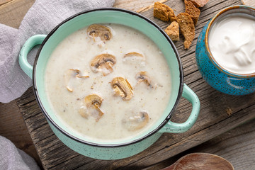 Turquoise ceramic pan with mushroom cream soup, cup with sour cream, crackers, grey napkin on old cracked wooden board.