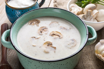 Turquoise ceramic pan with mushroom cream soup, cup with sour cream, fresh champignons on wooden background.