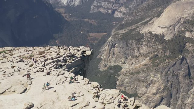 Aerial view of the flat top of Half Dome cliff brightly lit with the sun, with several tourists on it, walking near the edge. The steep stone slope of the mountain abruptly goes down. Yosemite. 4K