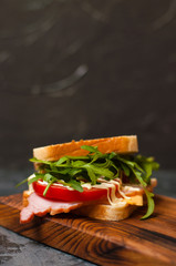 Very tasty big sandwich with ham or balyk, arugula, cheese, tomatoes and mayonnaise on a cutting board in rustic style on a gray background view from the side, copy space
