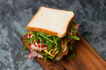 Huge delicious sandwich with ham and sturgeon, arugula, cheese, tomato and mayonnaise on a cutting board in a rustic style on the gray background view from the top