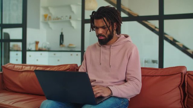 Creative african american freelance man with dreads working on business projecpt using laptop remotely at home office. Positive black hipster guy networking with laptop pc on sofa indoors.