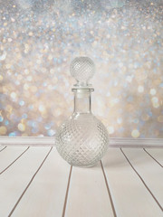 An empty round glass decanter on a glittering gold background with a bokeh effect on the wall and on the smooth beige boards of the table top. Free space for text.