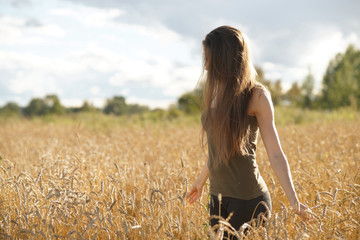 Fototapeta na wymiar Elegant woman with long hair standing waist-deep in wheat field and looking down. Hands touching beige ears of wheat