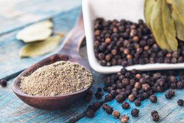 Ground black pepper in a spoon lies near the black peas scattered on the table and a bay leaf against the background of blue boards.