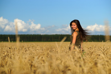 Beautiful woman with long hear standing waist-deep in wheat field, turning head backwards. Hands touching beige ears