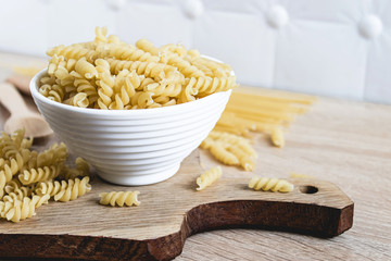 Dry pasta in a bowl on a cutting board on a light table background.