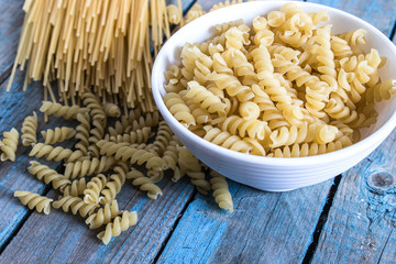 Dry pasta in a bowl on a background of blue boards near a pile of different pasta.