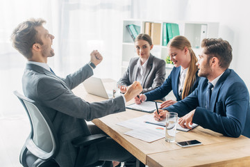 Side view of smiling employee showing yeah gesture near recruiters at table
